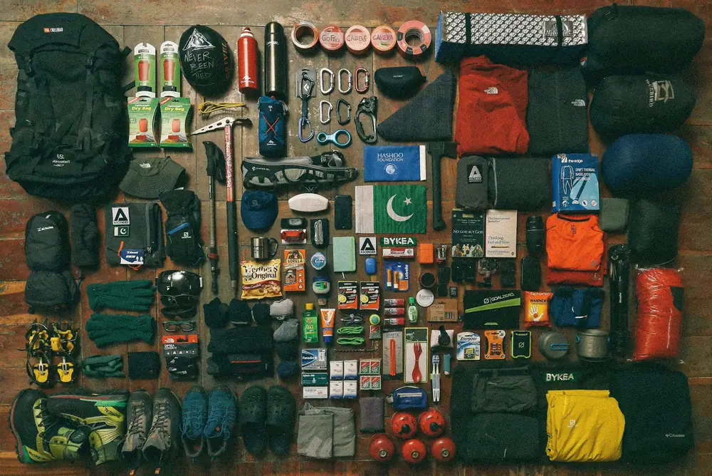 Outdoor gear laid out in neat rows on a dark mat, including packs, clothing, stoves and small accessories