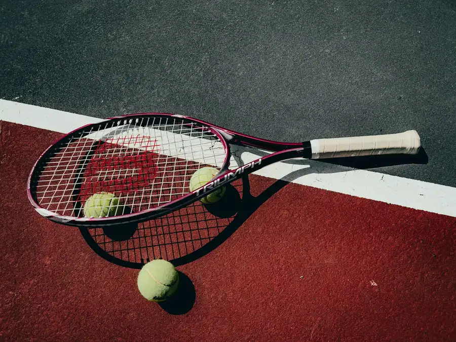 A tennis racket and three tennis balls resting on a red hard court