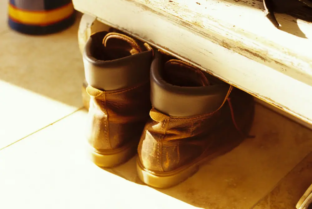 Worn leather boots tucked under a wooden bench in a sunlit hallway